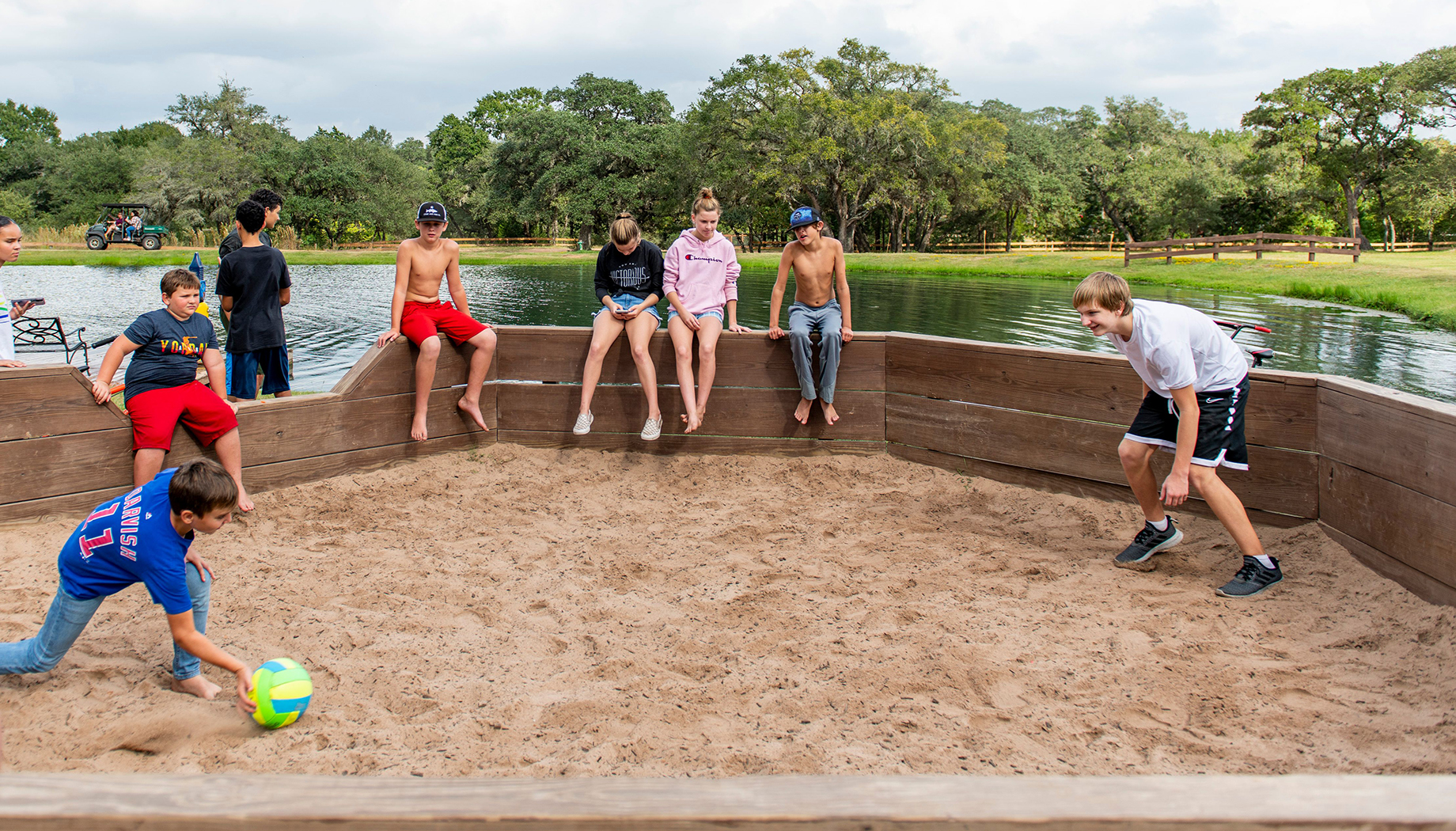 Two boys playing a game of Gaga Ball in a sand pit with people watching nearby.
